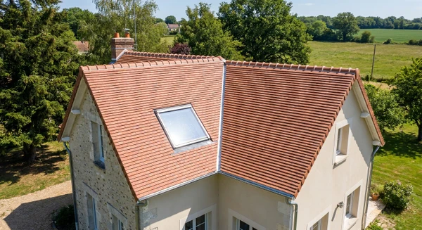 Photographie aérienne ou vue en légère plongée d'une maison individuelle avec toit en tuiles ou ardoises dans un contexte rural ou péri-urbain typique des Yvelines, montrant clairement la toiture en bon état ou en cours de rénovation. L'image doit capturer l'essence du métier de couvreur-zingueur en mettant l'accent sur les éléments de toiture (couverture, gouttières visibles, solins). Journée ensoleillée, ciel dégagé. Pas de personnes visibles. Composition professionnelle et nette.