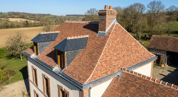 Photographie en vue aérienne ou légère plongée d'une maison individuelle traditionnelle aux toits en tuiles rouges ou ardoises grises, située dans un environnement rural ou péri-urbain des Yvelines. La maison doit être clairement visible avec ses éléments de toiture en détail : gouttières métalliques, solins, chéneaux bien visibles. Contexte paysager typique du Nord de l'Île-de-France avec végétation environnante. Jour ensoleillé, ciel dégagé. Aucune personne visible. Composition nette et professionnelle mettant l'accent sur la structure du toit et les éléments de zinguerie.