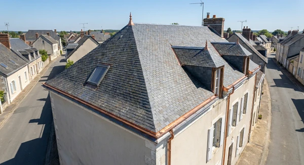 Photographie aérienne ou vue en légère plongée d'une maison ancienne typique de Nogent-le-Rotrou, avec toiture en ardoises grises ou tuiles, située dans un contexte urbain ou péri-urbain de province. La maison doit présenter clairement ses éléments de toiture : gouttières métalliques visibles, solins, chéneaux. Environnement architectural et paysager caractéristique de la région Perche. Jour ensoleillé, ciel dégagé. Aucune personne visible. Composition nette et professionnelle mettant en avant la structure du toit et les détails de zinguerie, distincte des maisons rurales isolées déjà sélectionnées.